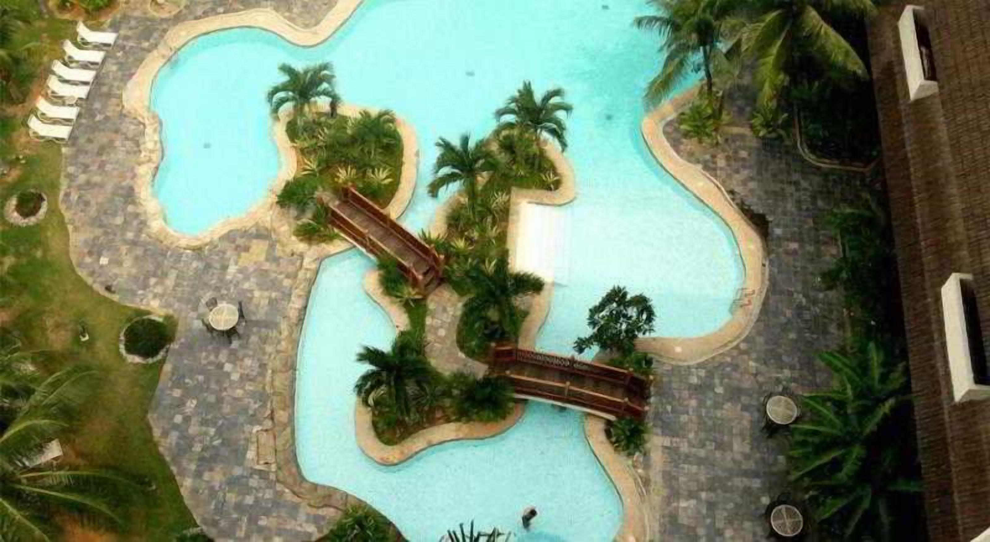 Piscina con un puente, rodeada de palmeras y un edificio.