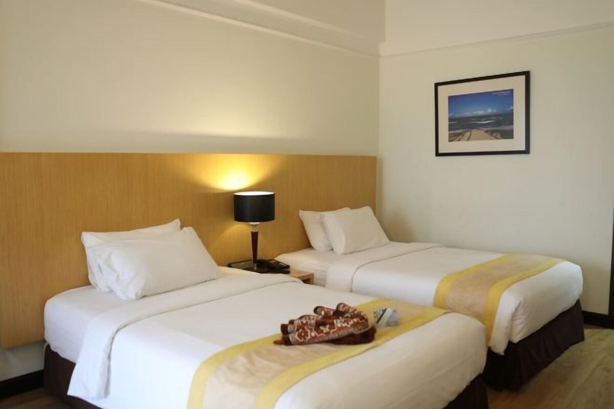 Quarto de hotel com duas camas, mesa de cabeceira com abajur, quadro na parede e bandeja com chinelos na cama.