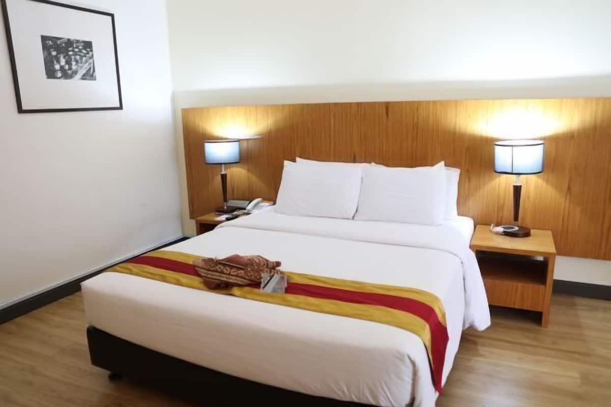 Une chambre d’hôtel avec un lit, deux tables de chevet avec des lampes, un tableau encadré au mur et une couverture à rayures rouges et jaunes sur le lit.