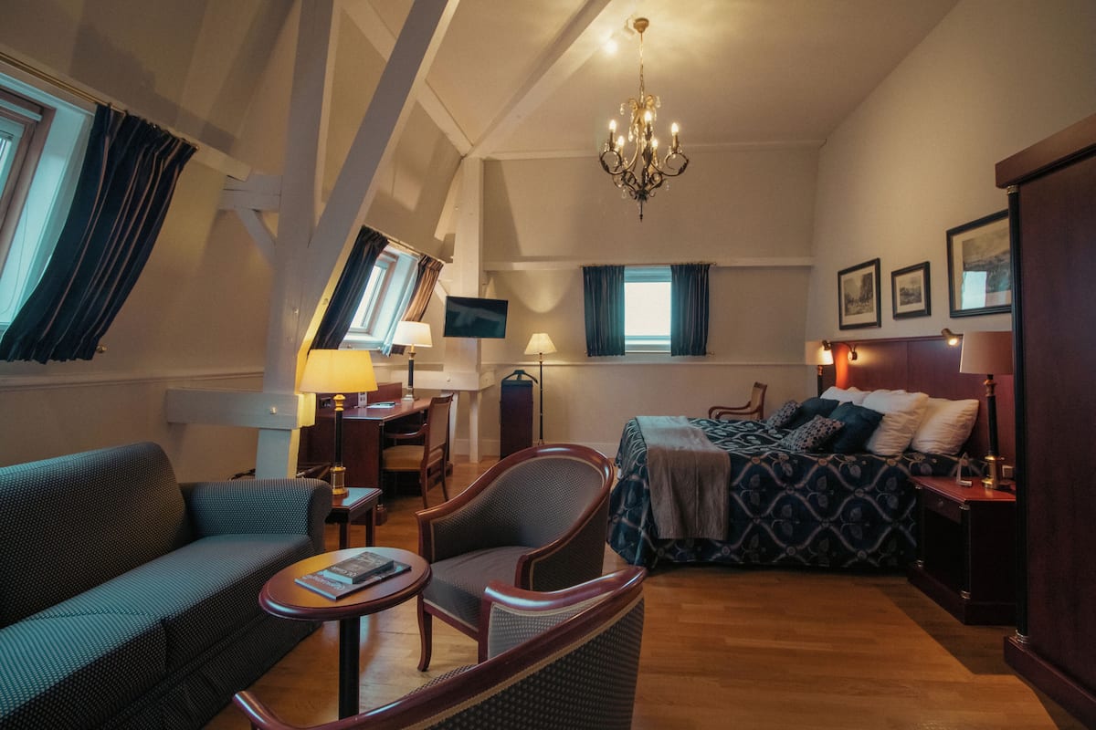 A hotel room with a bed, a sofa, a chair, a desk, a TV, and a chandelier.