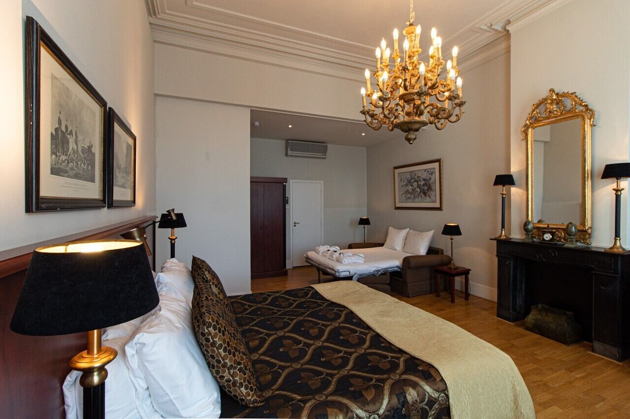 A hotel room with a large bed, a chandelier, a fireplace, and framed pictures on the wall.