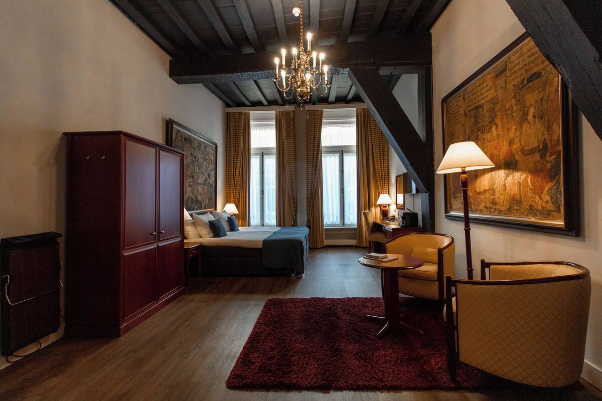 A bedroom with a bed, a desk, a chair, a red rug, a chandelier, and a large painting on the wall.