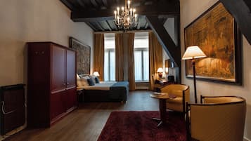 A bedroom with a bed, a desk, a chair, a red rug, a chandelier, and a large painting on the wall.