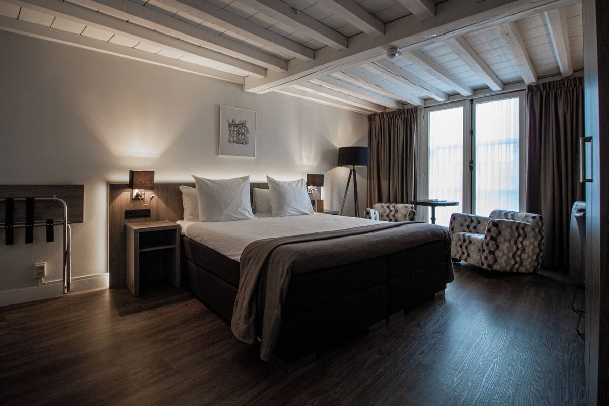 A modern hotel room with a large bed, bedside tables, a desk, a chair, and a view of the outdoors.