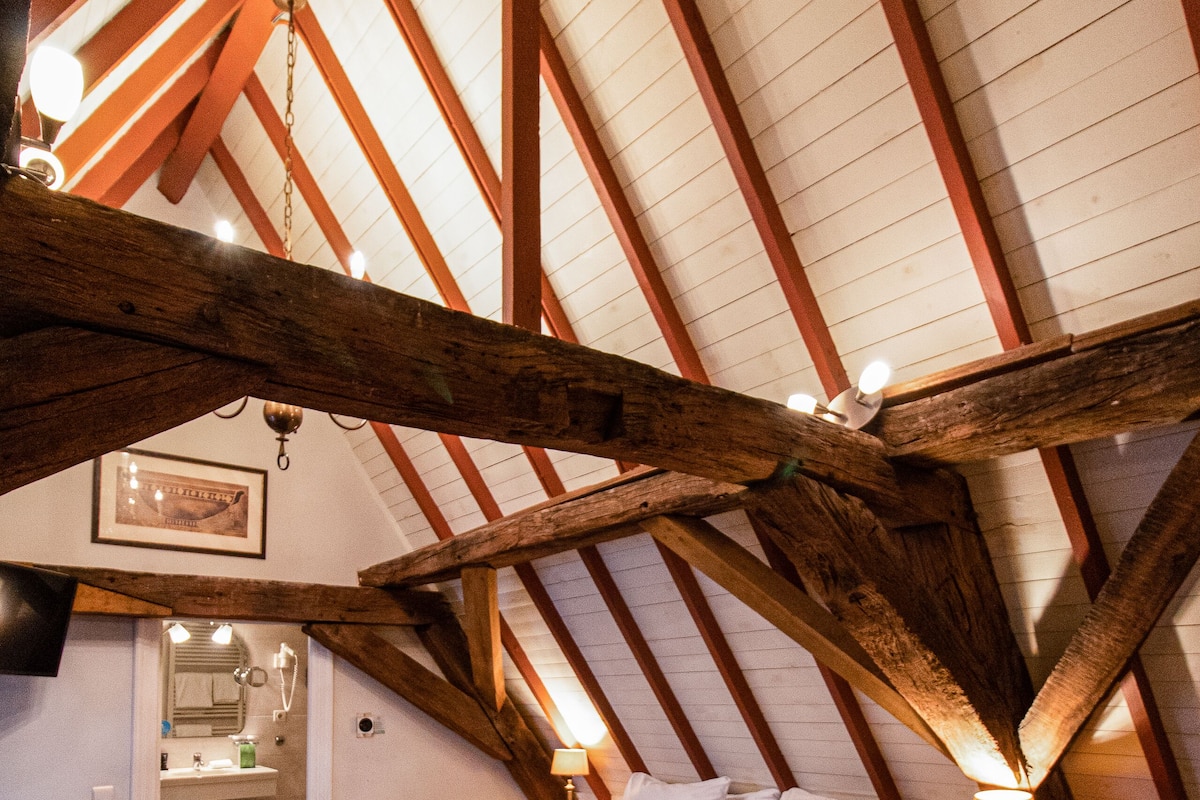 A bedroom with a sloped ceiling, wooden beams, a bed with pillows, a bedside table, and a nightstand.