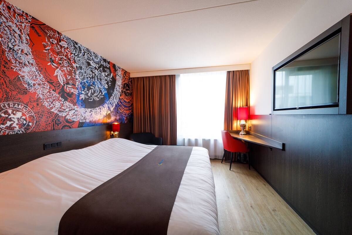 A hotel room with a large bed, a desk with a chair, a TV mounted on the wall, and a patterned wall.