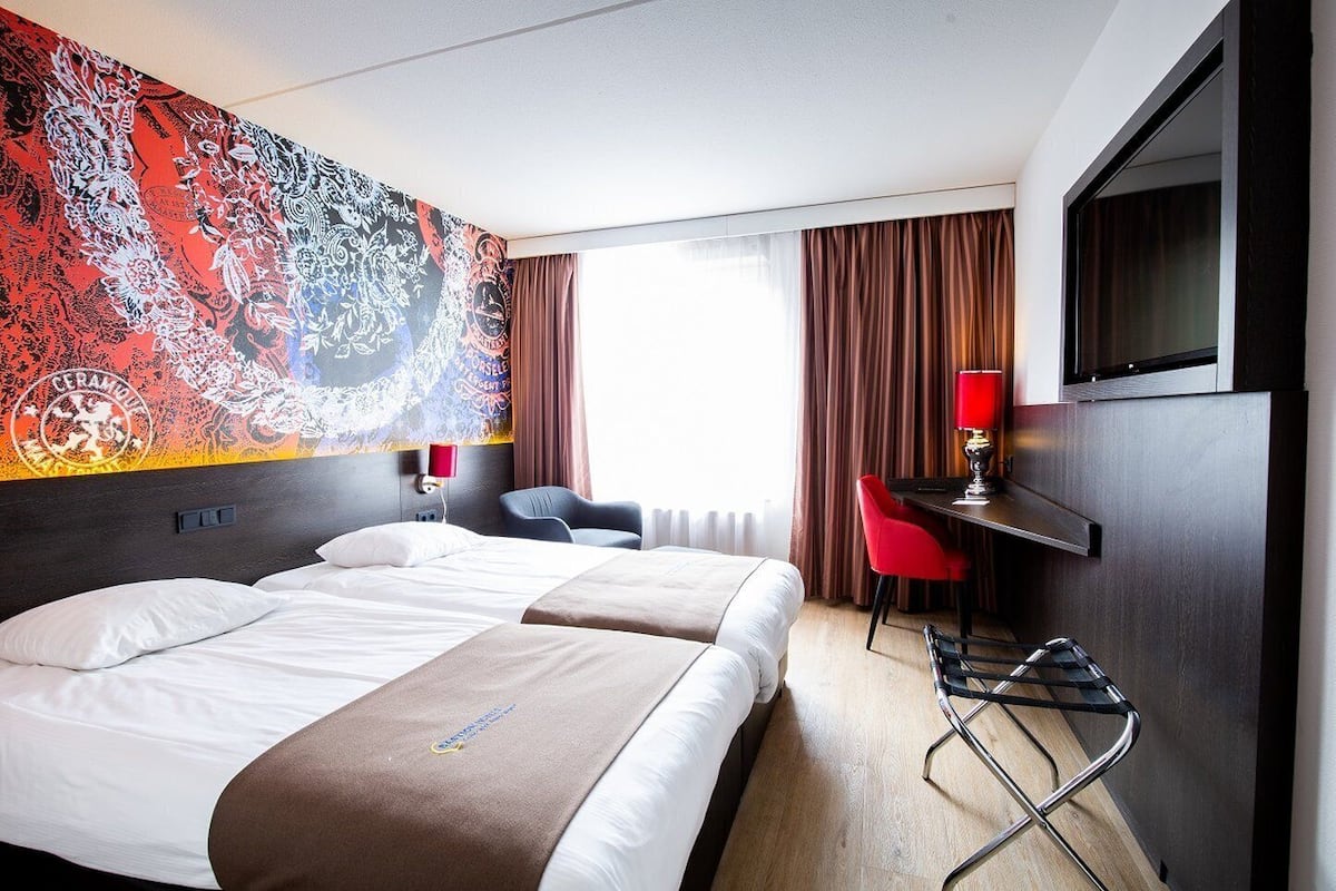 A hotel room with two beds, a desk, a chair, a TV, and a large abstract wall mural.