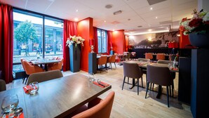 A restaurant with a modern interior, featuring wooden tables and chairs, red curtains, and large windows offering a view of the outside. - Bastion Hotel Maastricht Centrum (Maastricht)