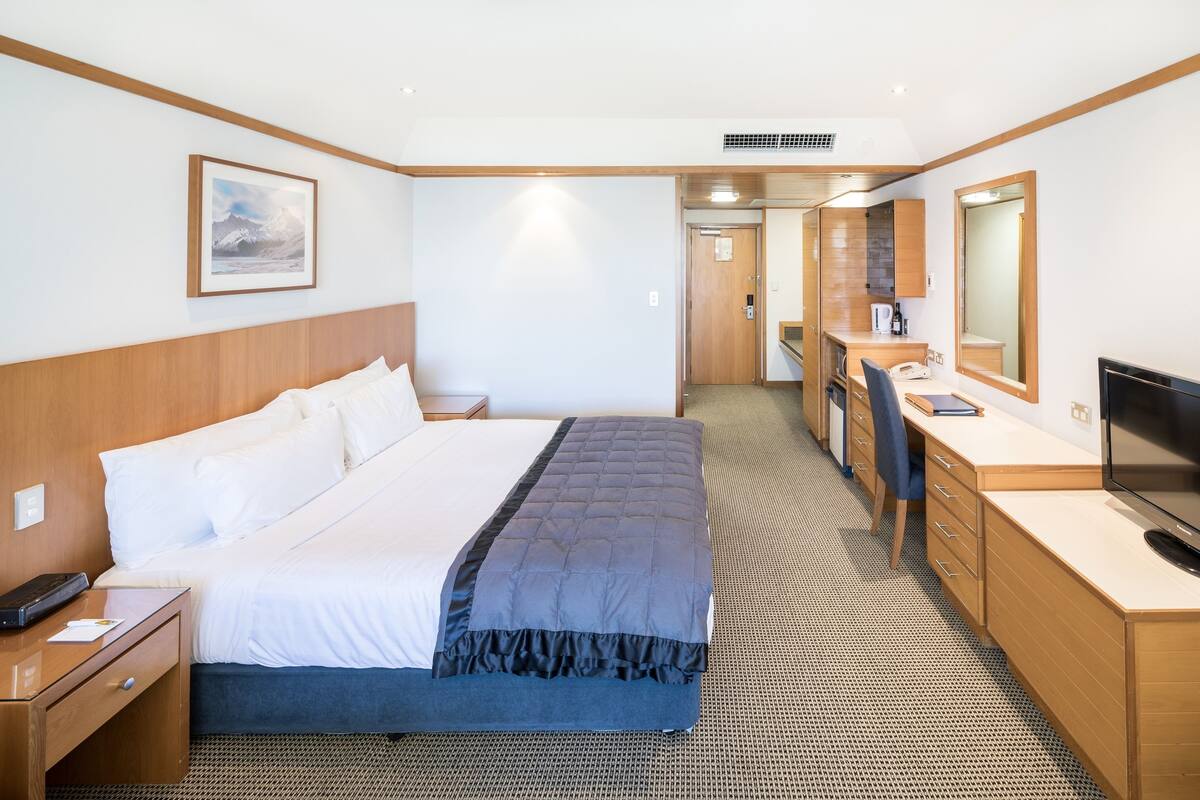 A hotel room with a bed, bedside table, desk, chair, television, and a bathroom visible through an open door.