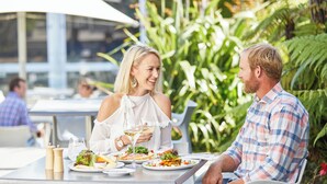 A couple dining at an outdoor restaurant with a view of a modern building and lush greenery in the background. - Commodore Airport Hotel (Christchurch)