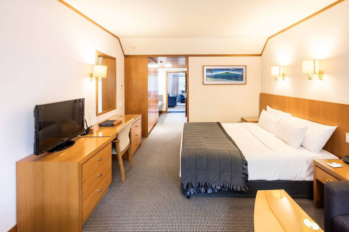 A hotel room with a bed, a desk with a TV, a chair, a bedside table, and a picture on the wall.