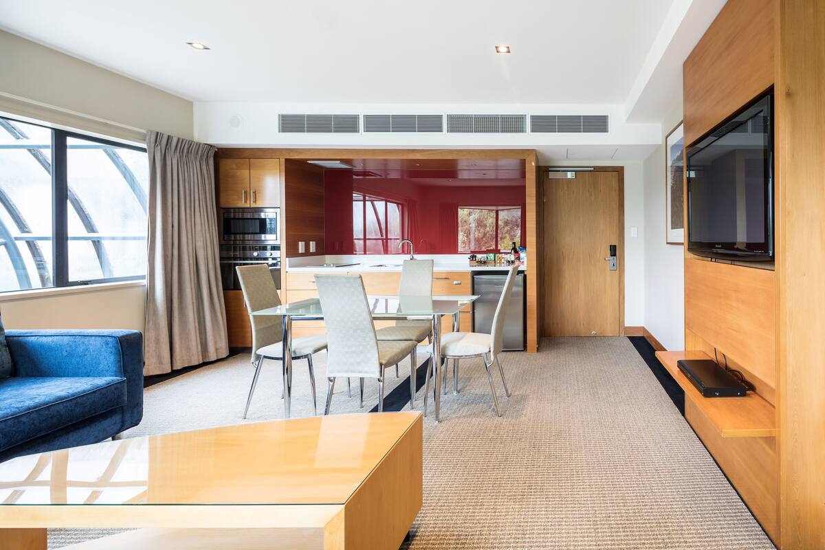 A modern hotel room with a dining area, kitchenette, and a large TV.