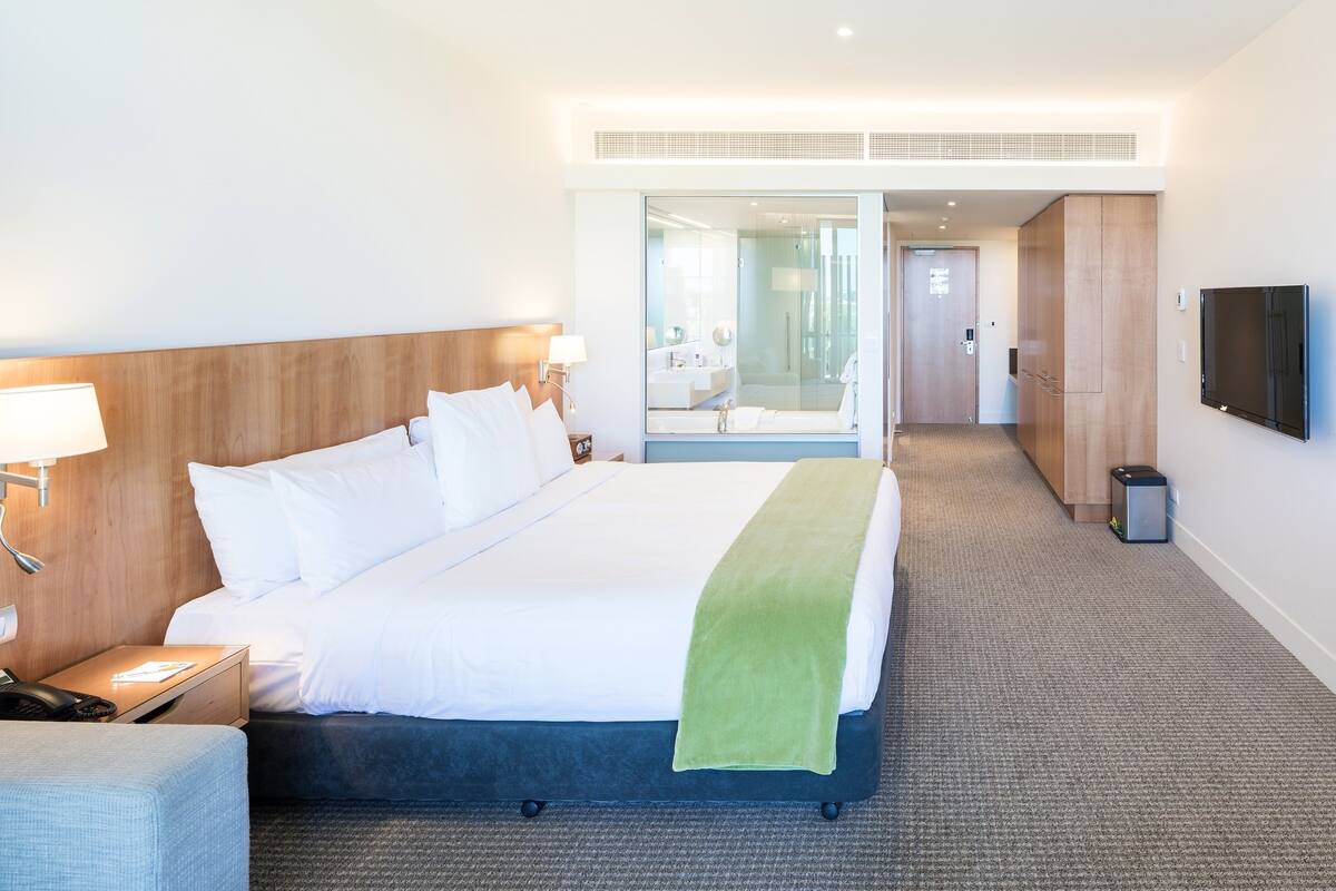 A modern hotel room with a large bed, a bedside table with a lamp, a TV mounted on the wall, and a view into the bathroom through a glass door.