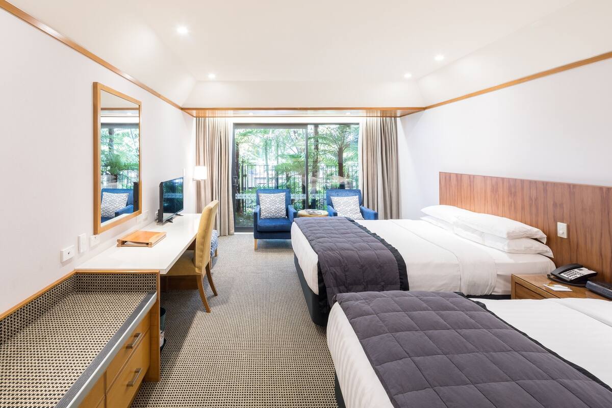 A hotel room with two beds, a desk, a chair, a TV, and a view of the outdoors.
