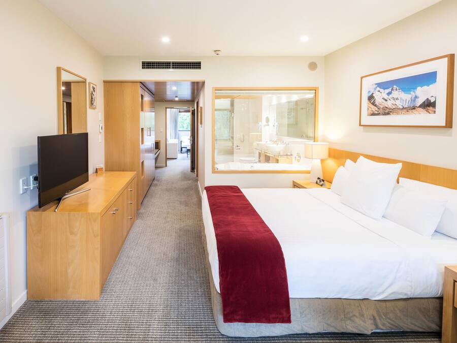 A hotel room with a bed, a TV, a desk, and a bathroom visible through a glass door.