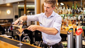 A bartender pouring a drink into a martini glass at a bar with a variety of liquor bottles in the background. - Commodore Airport Hotel (Christchurch)