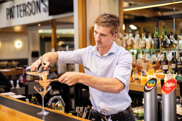 A bartender pouring a drink into a martini glass at a bar with a variety of liquor bottles in the background. - Commodore Airport Hotel (Christchurch)
