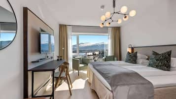 Ein modernes Hotelzimmer mit einem großen Bett, einem Schreibtisch mit Fernseher, einem Sessel und Blick auf einen verschneiten Berg durch große Fenster.