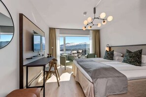 A modern hotel room with a large bed, a desk with a flat-screen TV, a chair, and a view of a snowy mountain through large windows. - Scandic Parken (Ålesund)
