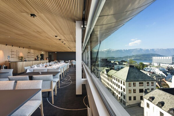 Un restaurante moderno con techo alto, paneles de madera y vistas a un paisaje urbano y montañas.