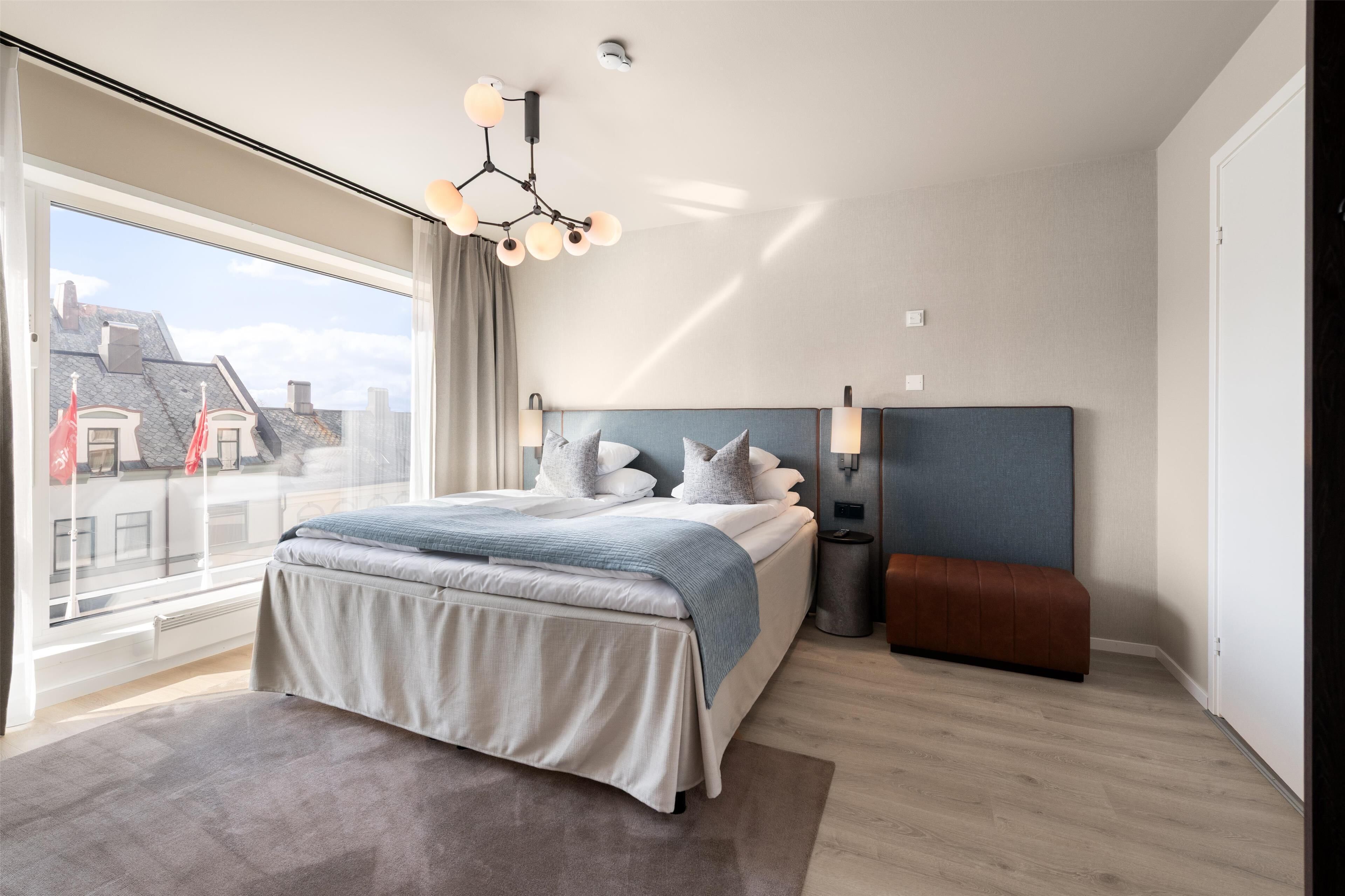 A modern bedroom with a large window, a bed with a blue headboard, a bedside table, and a bench.