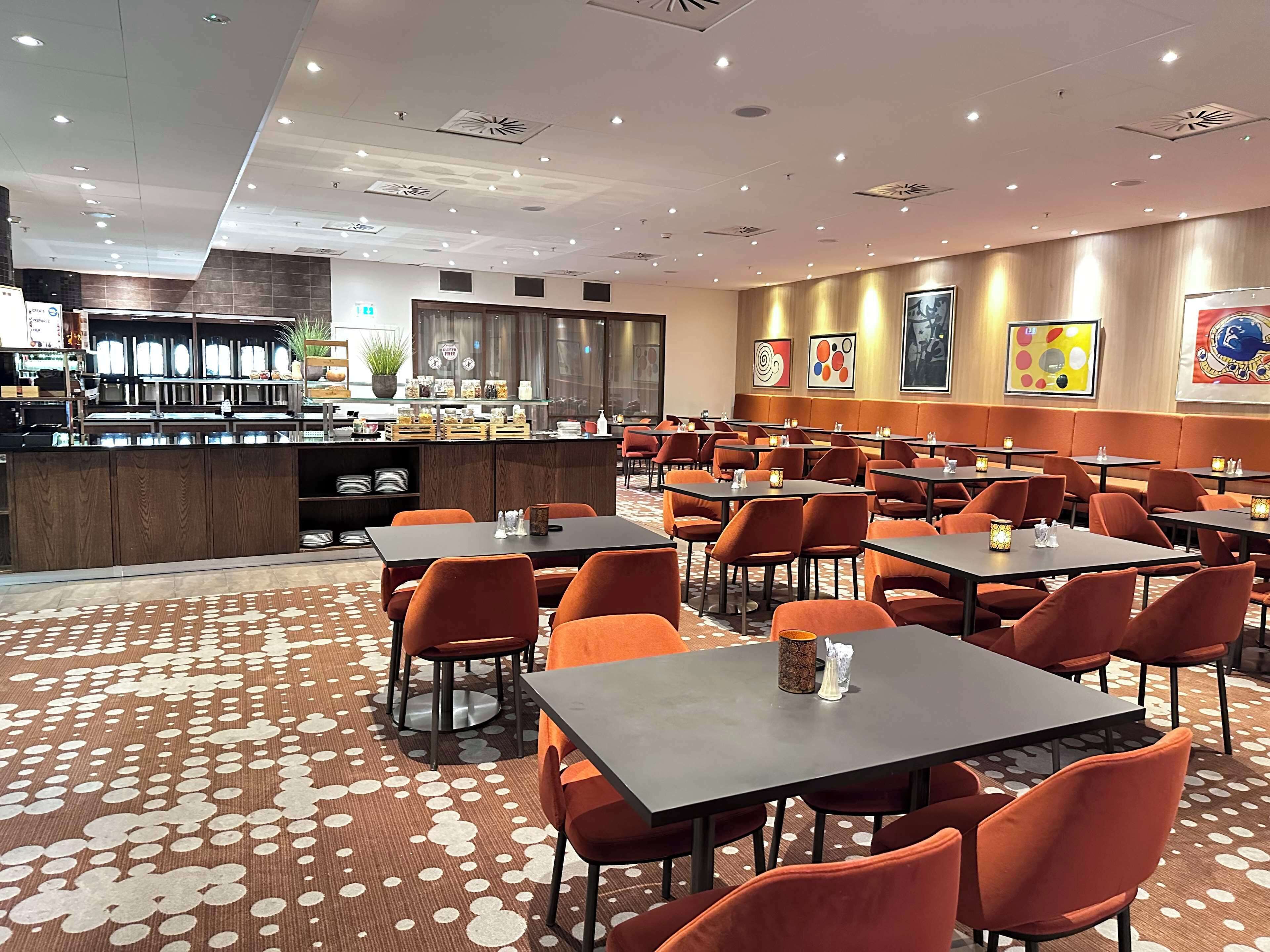 Un restaurante amplio con varias mesas y sillas anaranjadas, una barra con diferentes productos alimenticios y una alfombra estampada.