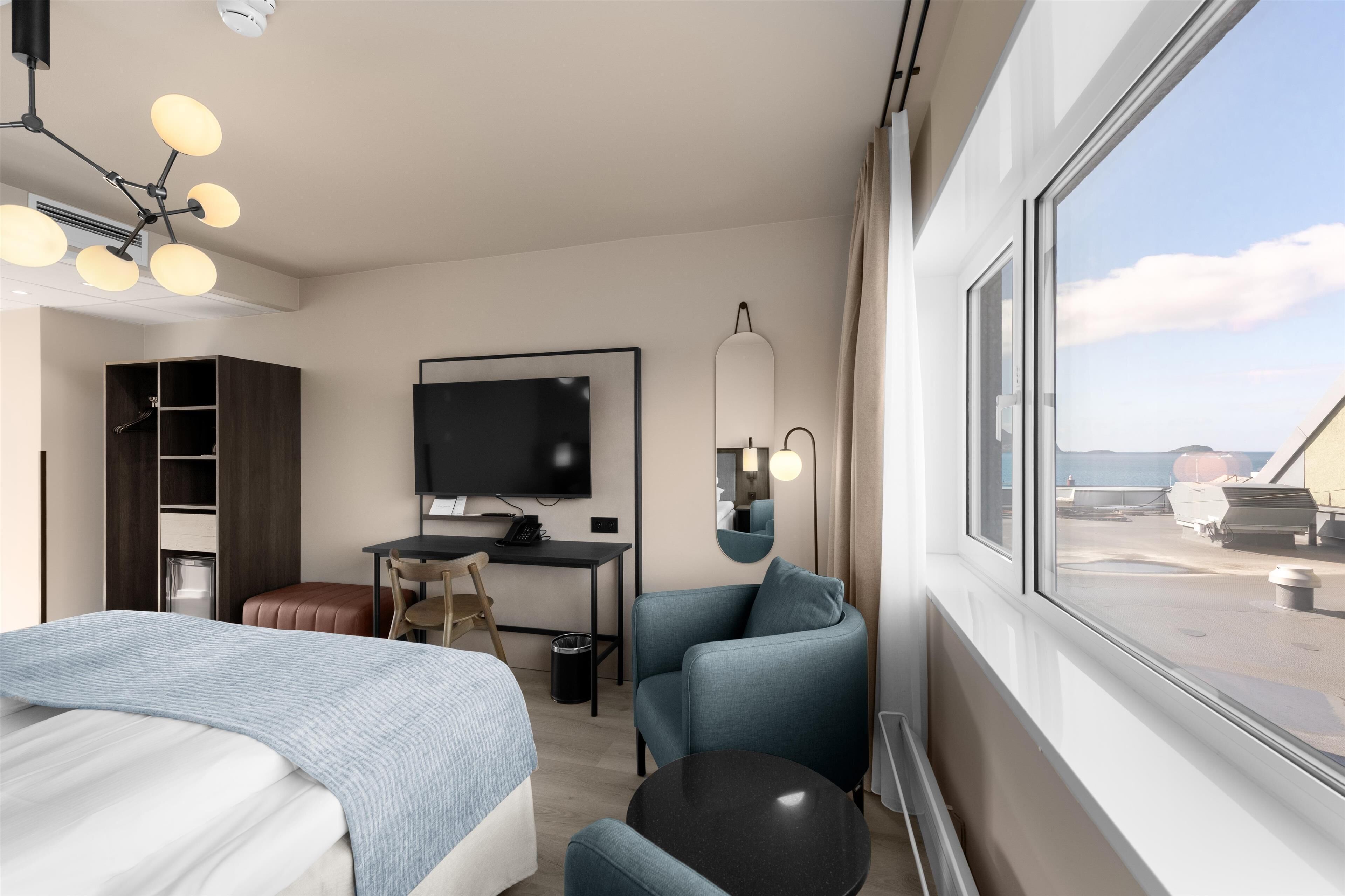 A modern hotel room with a bed, a desk, a chair, a TV, and a large window offering a view of the sea.