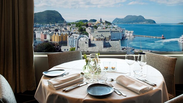 Une table à manger dressée avec des assiettes, des couverts et des verres, donnant sur une ville côtière avec ses bâtiments et son phare.