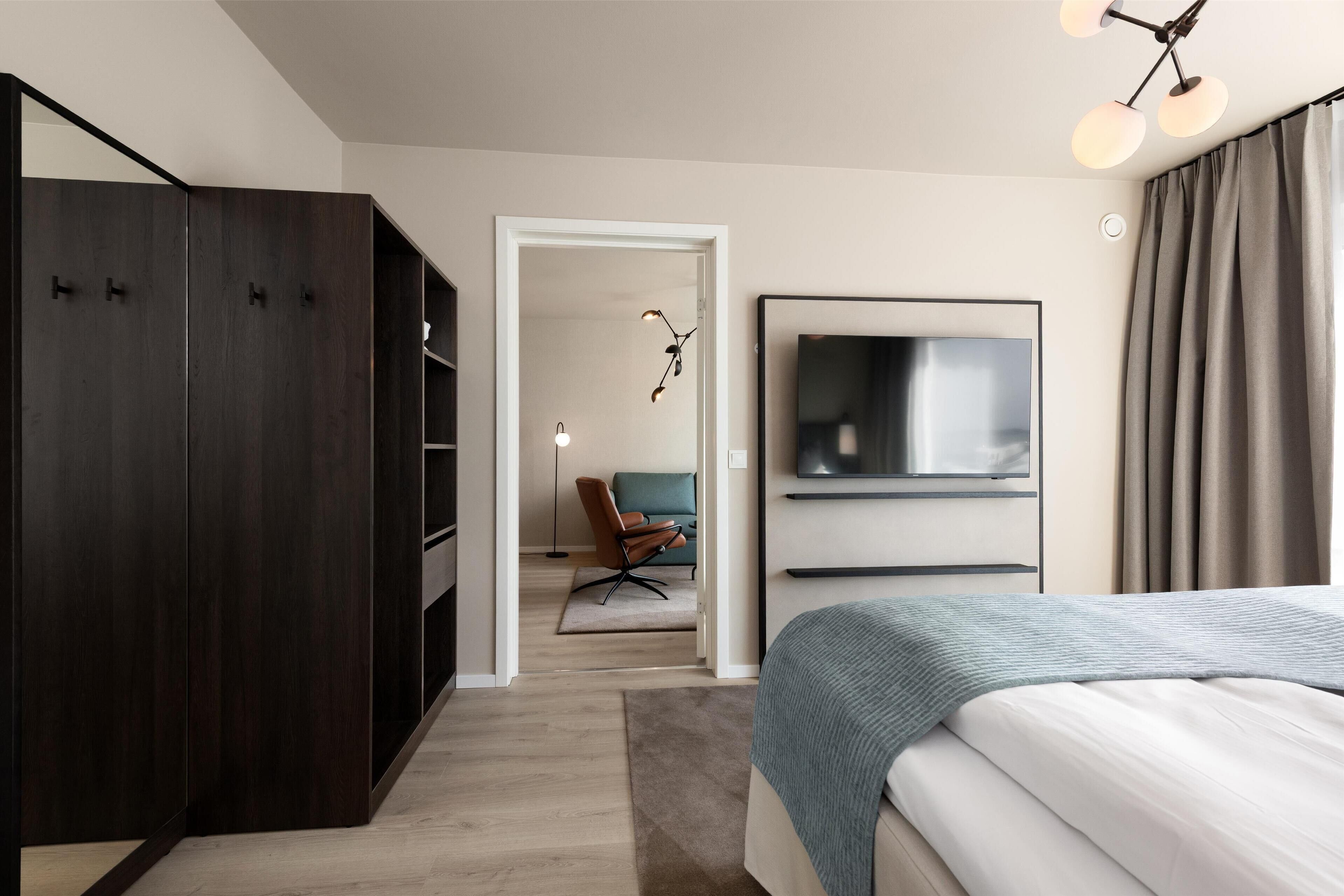 A modern bedroom with a large bed, a wardrobe, a TV, and a chair in the background.