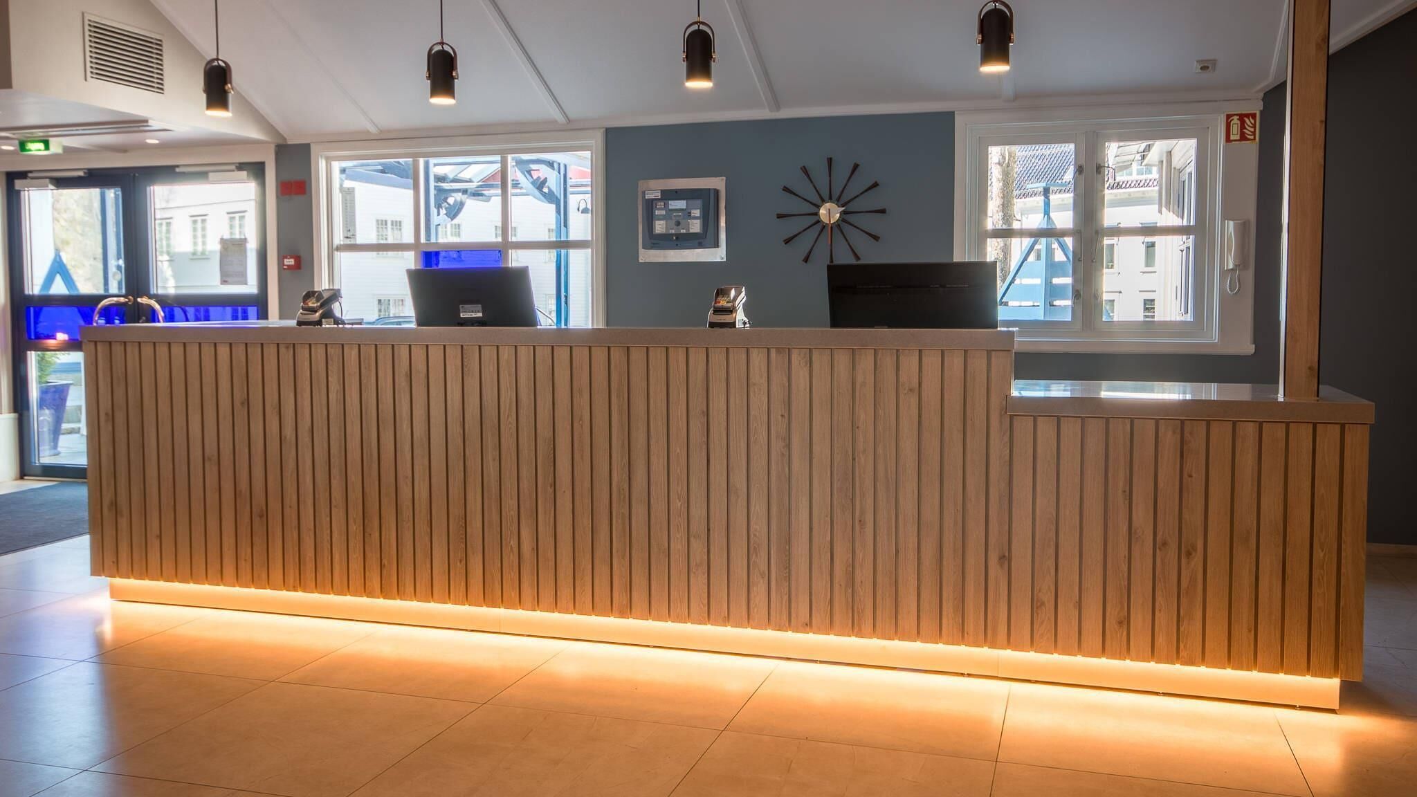 A reception desk with a wooden finish and integrated lighting, a blue wall clock, and a window with a view of buildings outside.