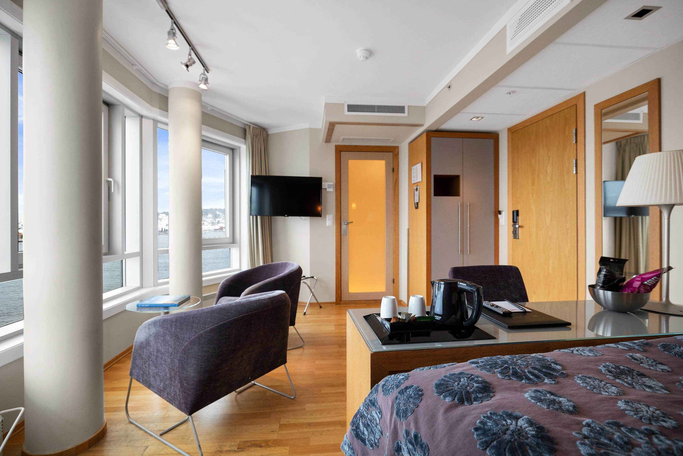 A hotel room with a bed, two chairs, a desk with a laptop, a television, and a window with a view.