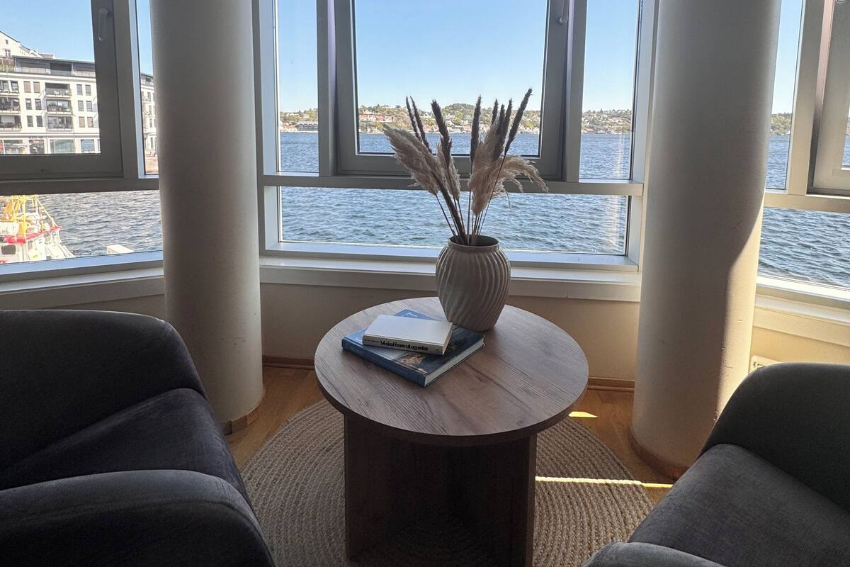 Een moderne woonkamer met een groot raam dat uitkijkt over een waterpartij, een ronde houten tafel met boeken en een vaas, en twee donkere fauteuils.