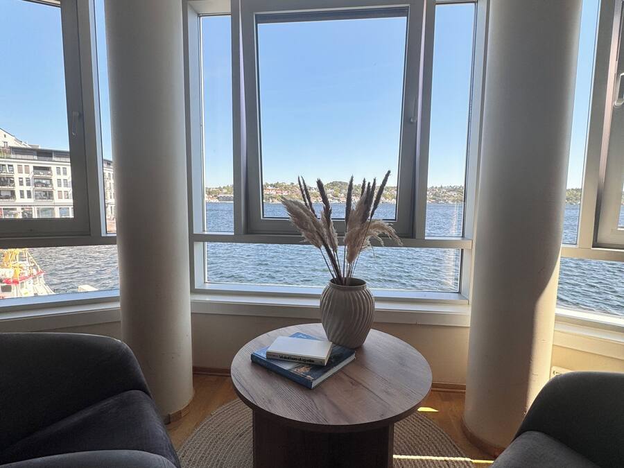 Una sala moderna con un ventanal grande con vista a una masa de agua, una mesa redonda de madera con libros y un florero, y dos sillones oscuros.