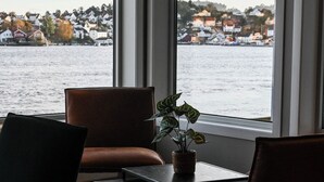 Quarto com uma grande janela que oferece vista para um corpo de água e uma área residencial. Há duas cadeiras e uma pequena mesa com uma planta.