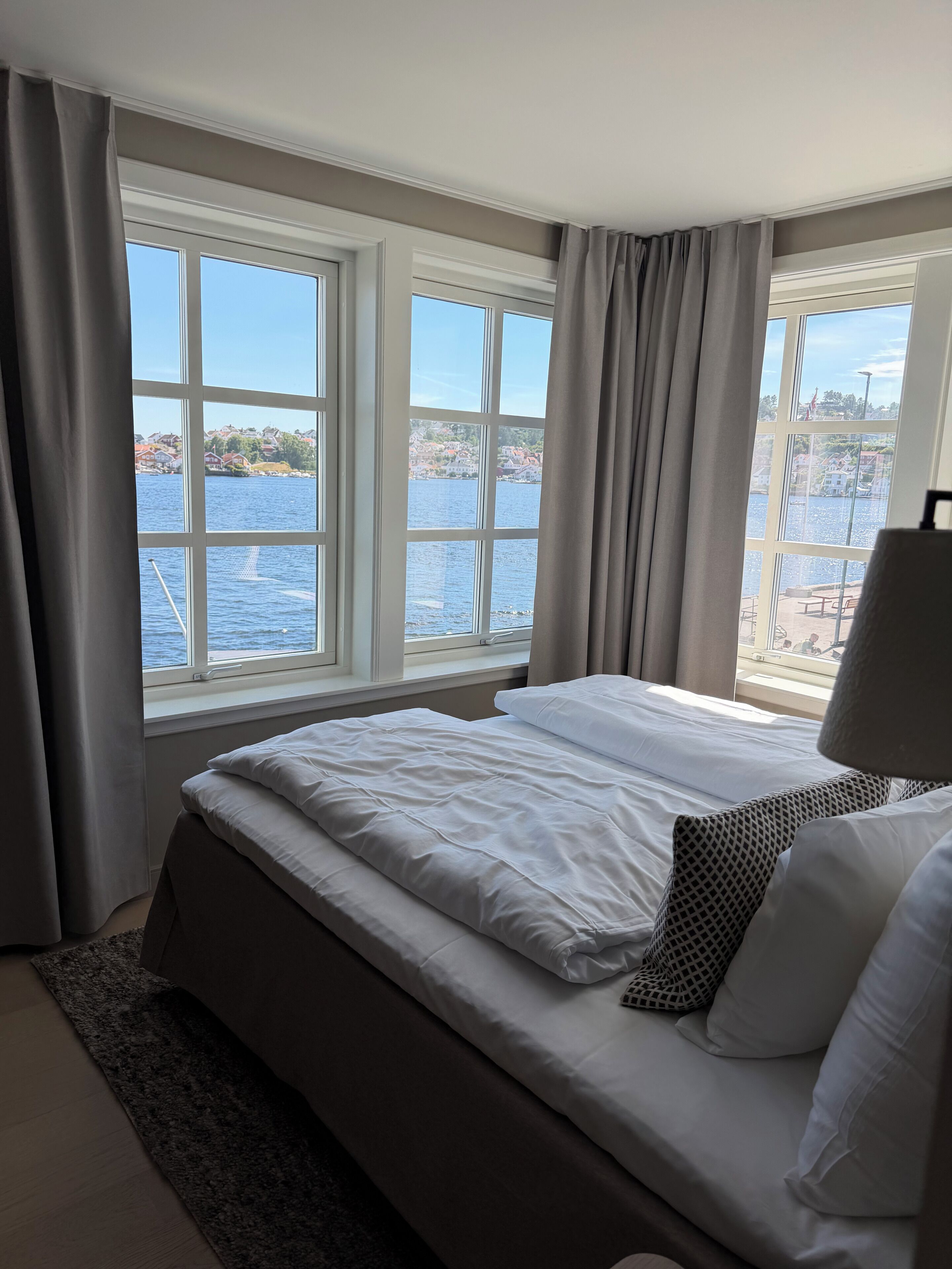 Un dormitorio con una cama grande, dos ventanas con cortinas, vista a un cuerpo de agua y una lámpara.