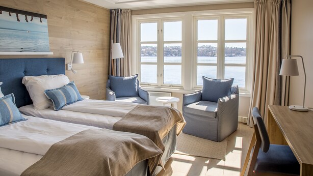 Quarto de hotel com duas camas, área de estar com uma cadeira e uma mesa, e uma janela grande com vista para a água.