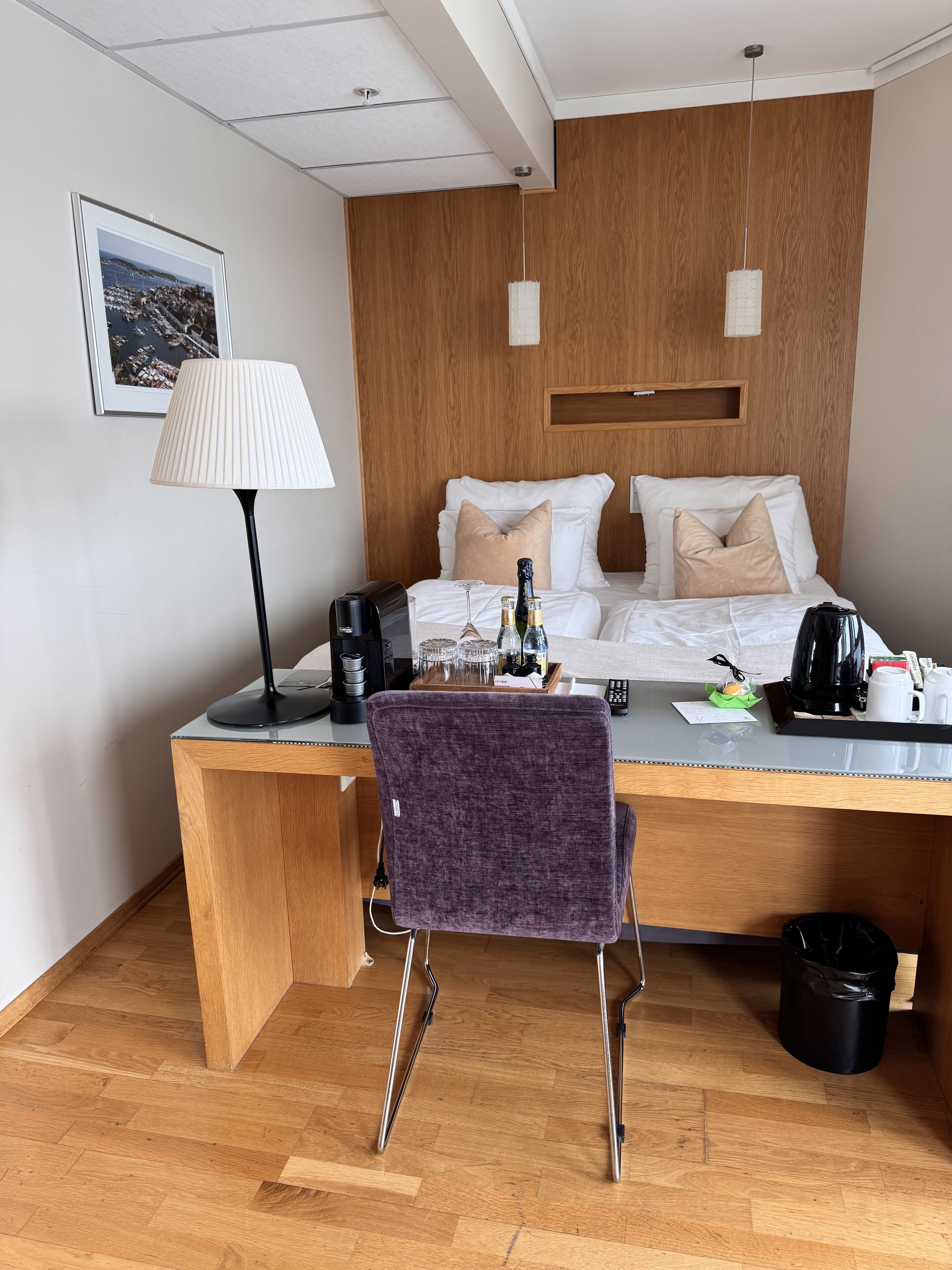 Una habitación de hotel con cabecera de madera, cama con ropa blanca, escritorio con lámpara, una silla y cafetera.