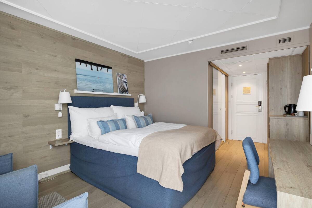 Een moderne hotelkamer met een groot bed, bedlampen, een houten hoofdbord, een klein bureau en uitzicht op de zee door een raam.
