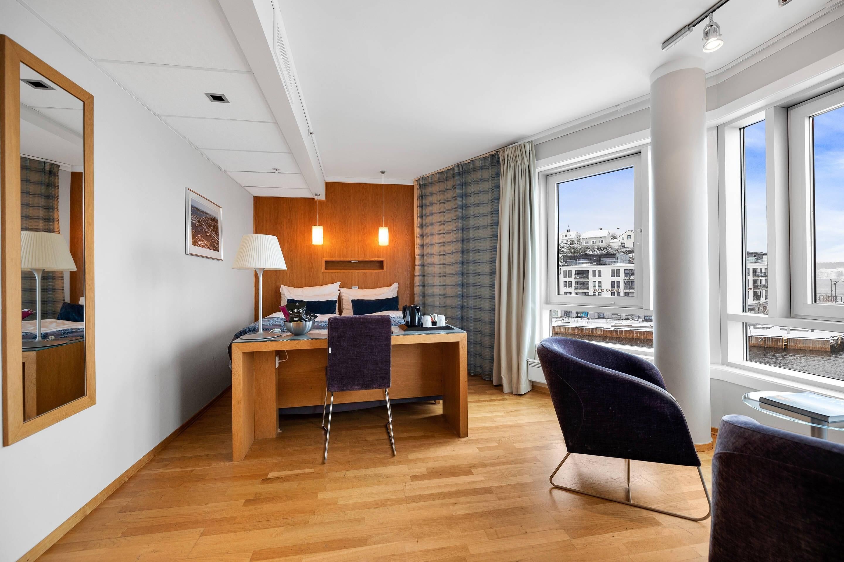 A hotel room with a bed, desk, chair, large windows, and a view of buildings.