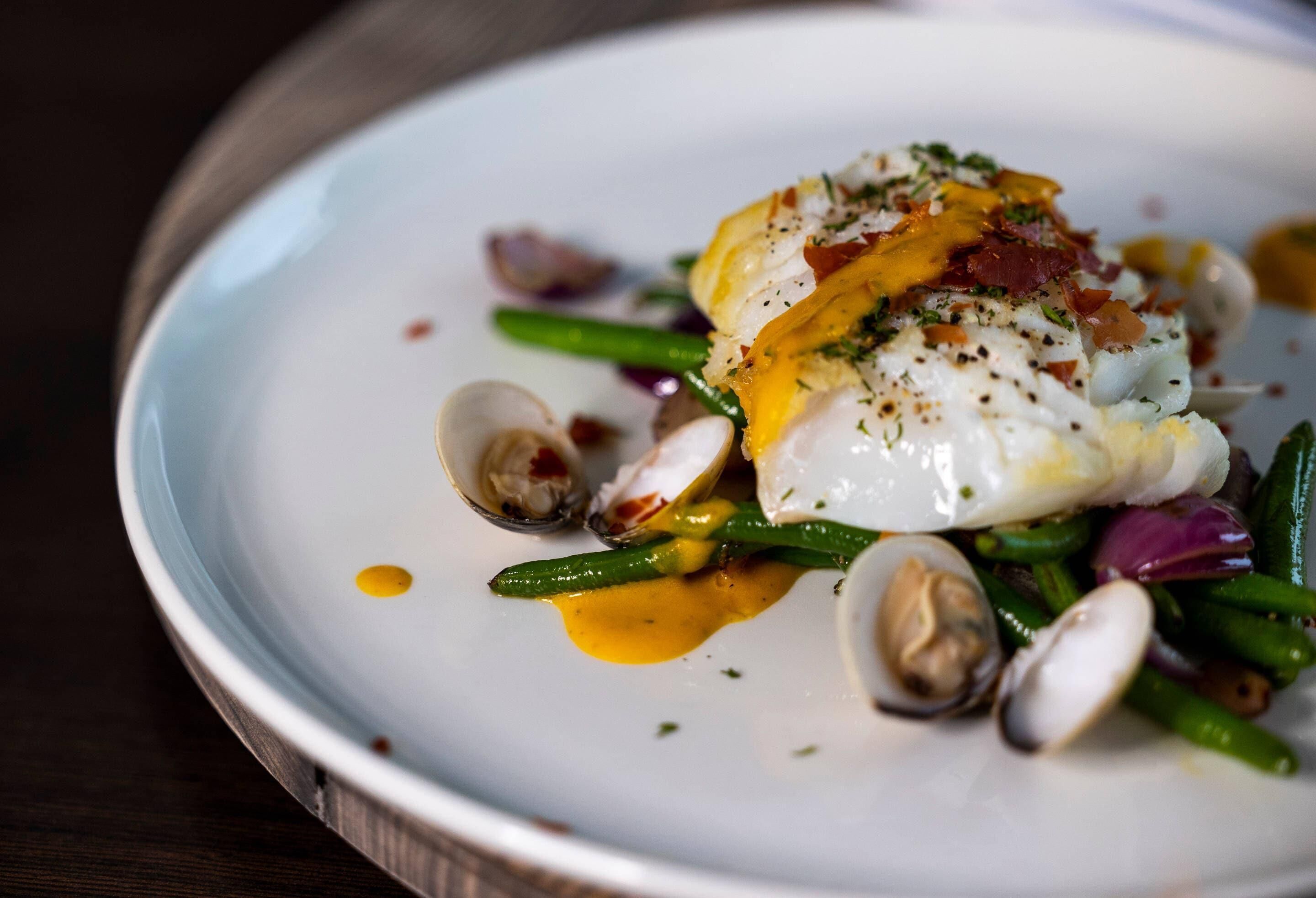 A white plate with a poached egg, green beans, clams, and a drizzle of sauce.