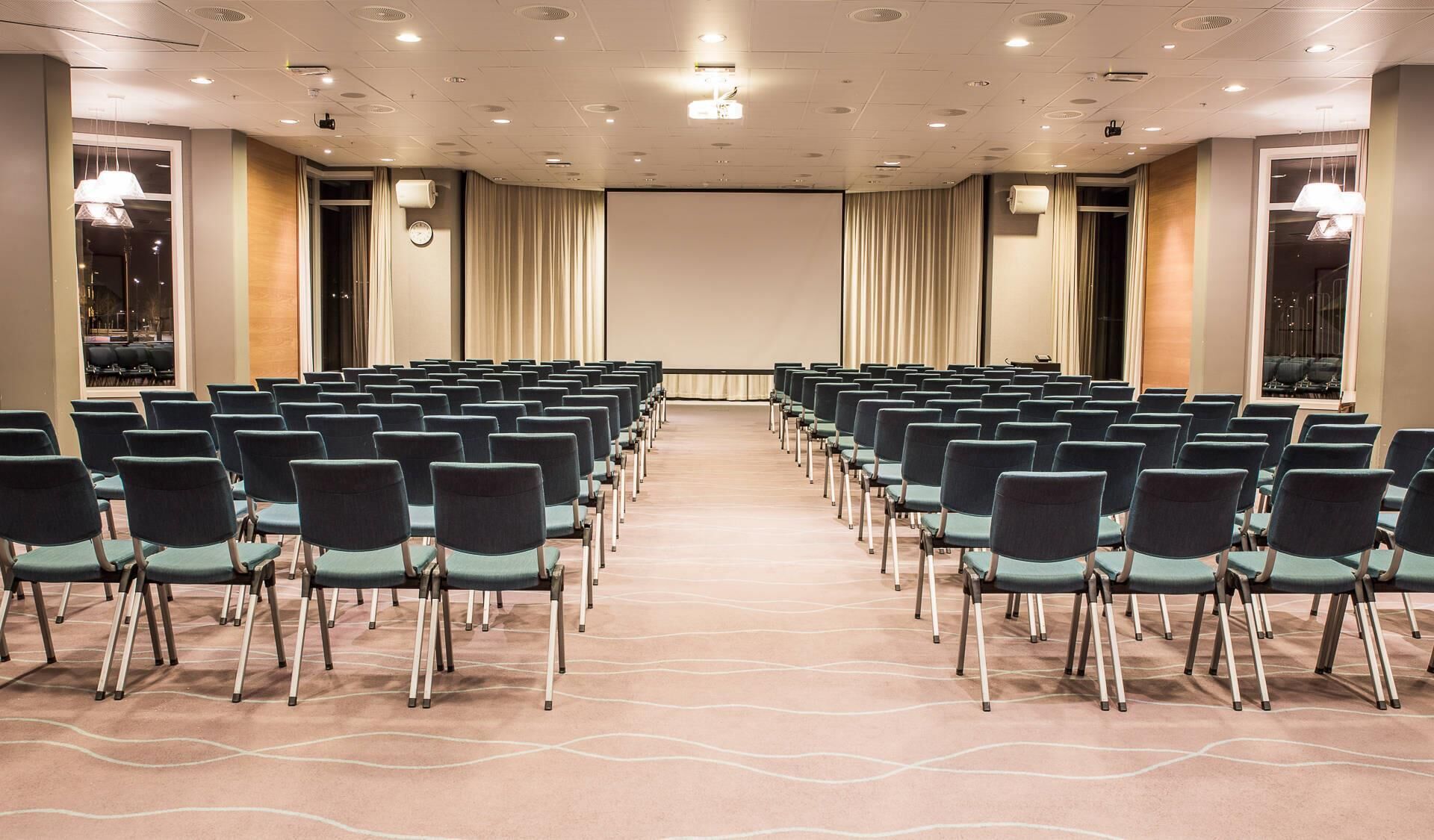 Un salón de conferencias con filas de sillas, una pantalla grande y un proyector.