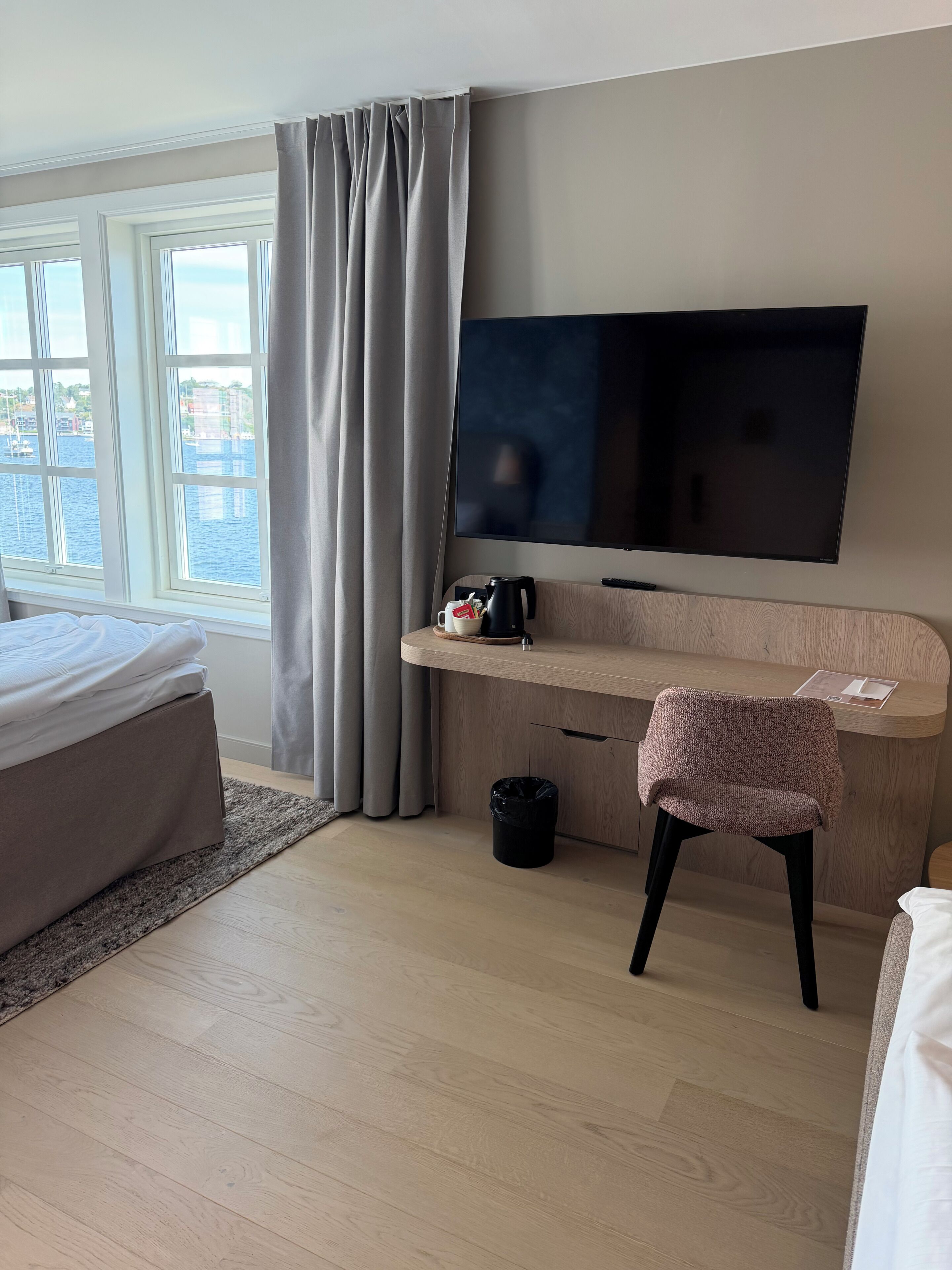 A hotel room with a bed, a desk with a TV, a chair, a coffee maker, and a view of the sea through large windows.