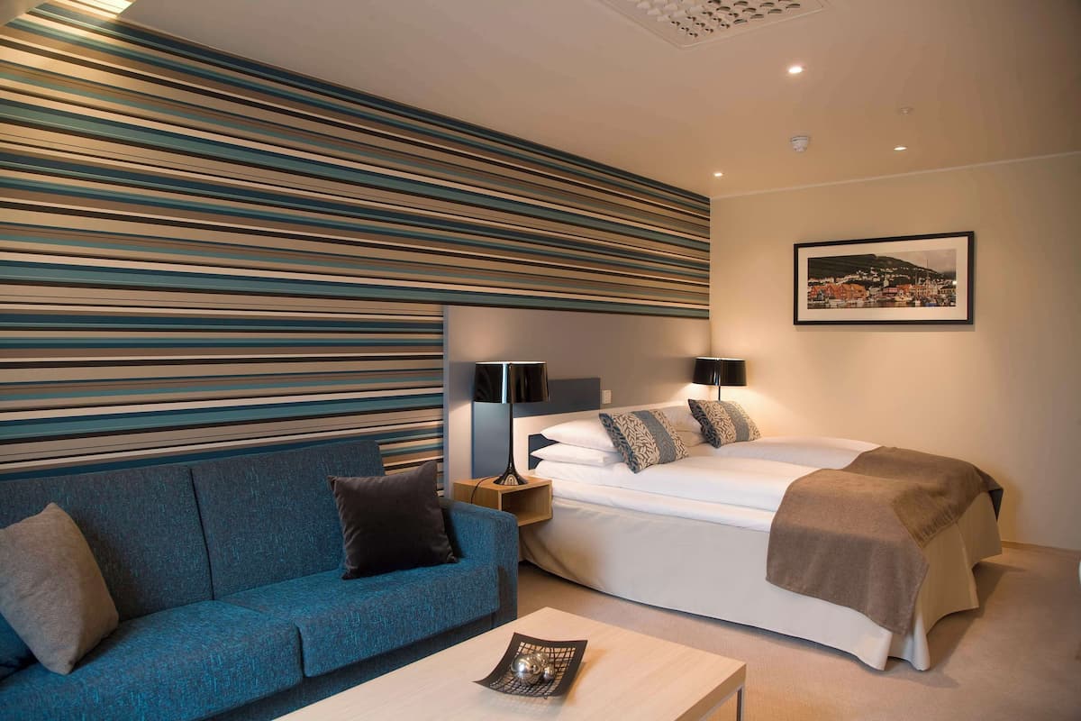 A modern hotel room with a sofa, a bed, a nightstand, and a lamp.