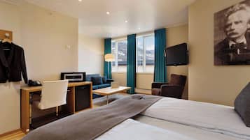 A hotel room with a bed, a desk with a laptop, a chair, a TV, and a window with curtains.