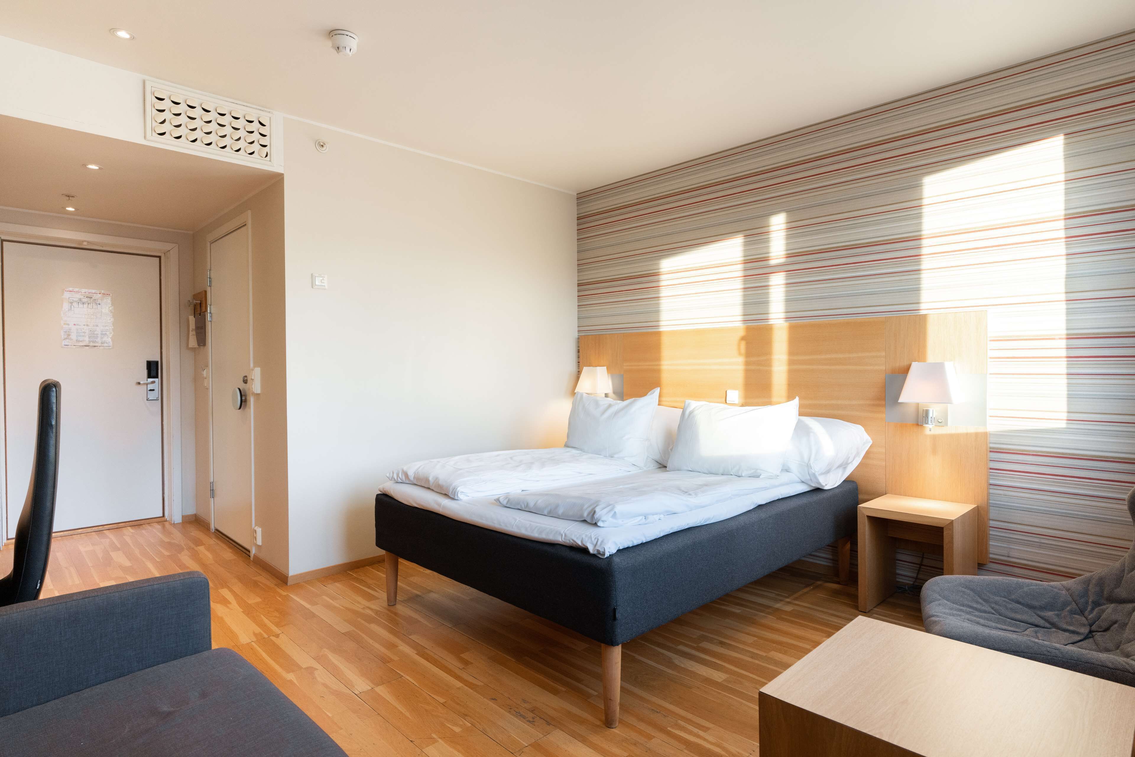 Una habitación de hotel moderna con una cama grande, una mesita de noche pequeña, una silla y suelo de madera.