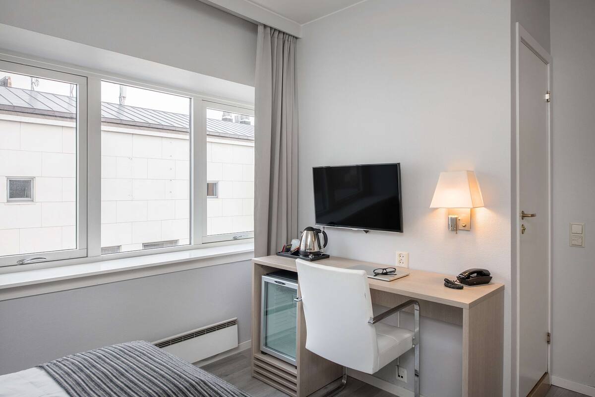 A hotel room with a bed, a desk with a TV, a lamp, a telephone, and a minibar.