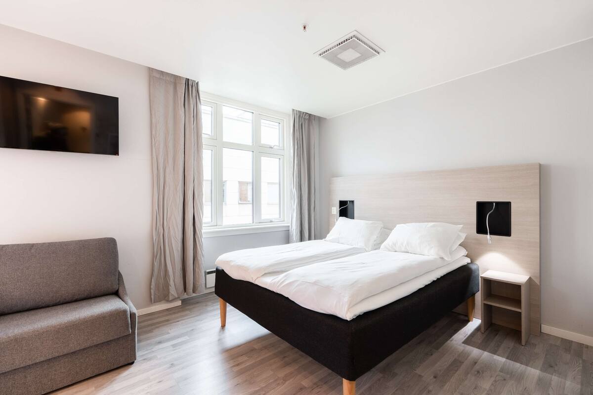 A modern bedroom with a bed, a grey sofa, a bedside table, and a large window with curtains.