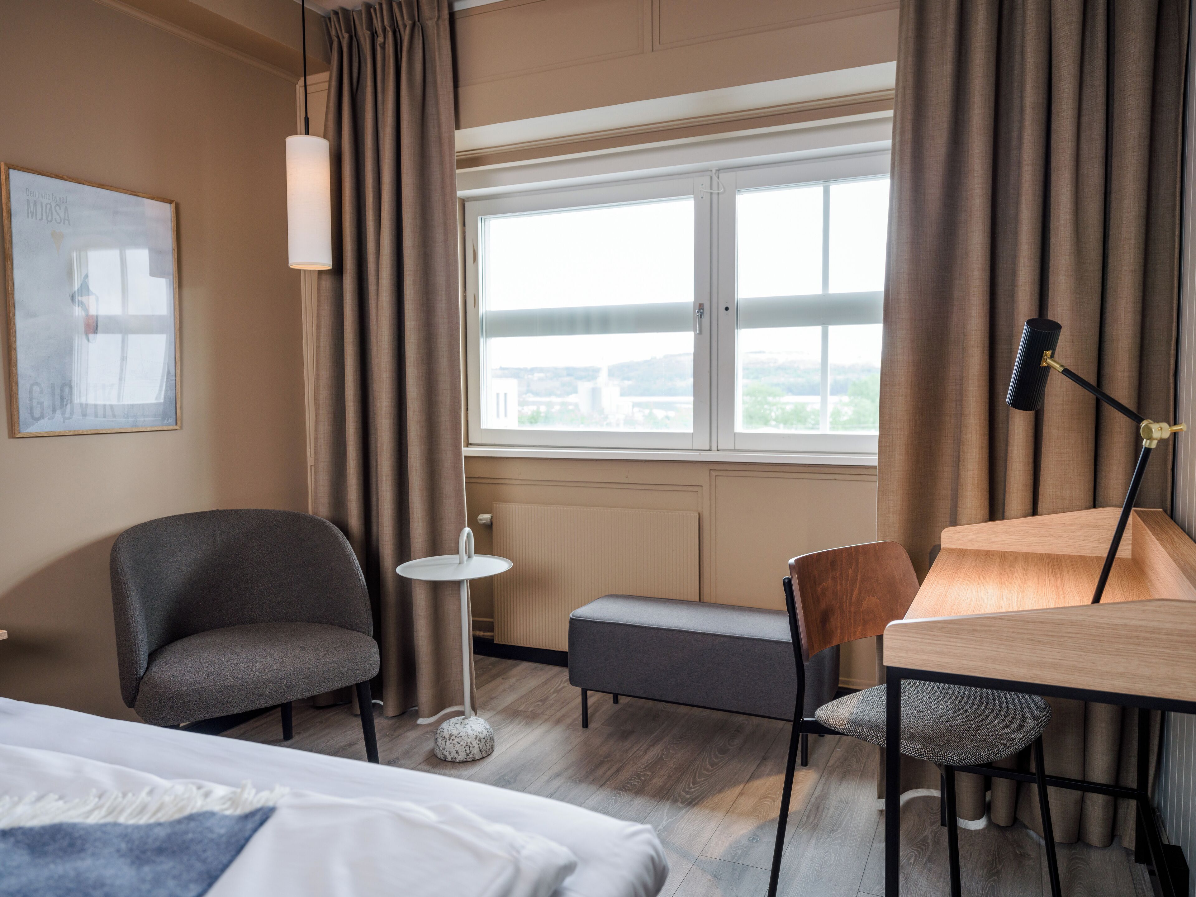 A hotel room with a bed, a desk, a chair, a small table, and a view of the city through the window.
