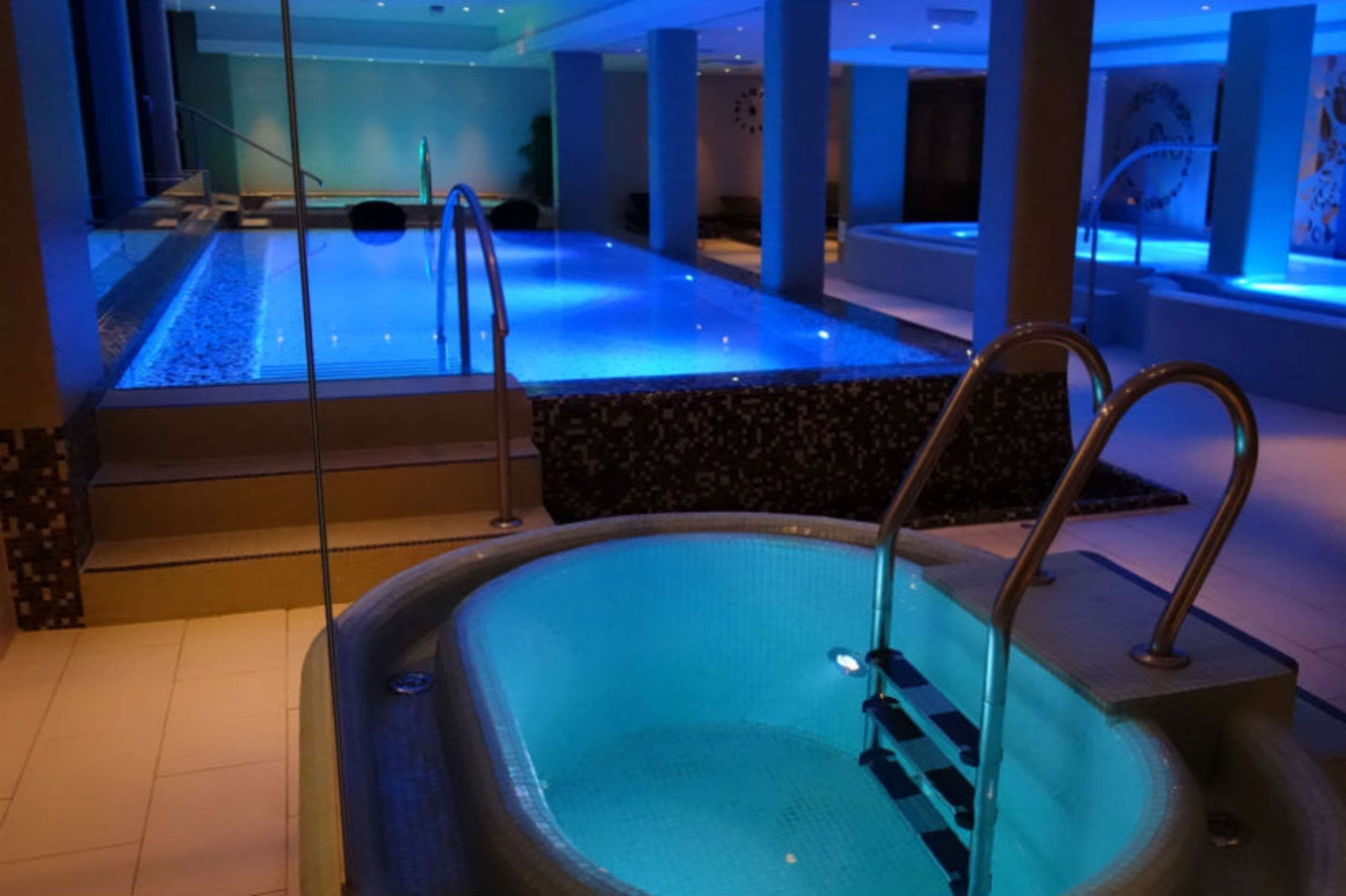 Una amplia piscina interior con una fuente iluminada en azul, rodeada de un suelo de baldosas y una pared de piedra.