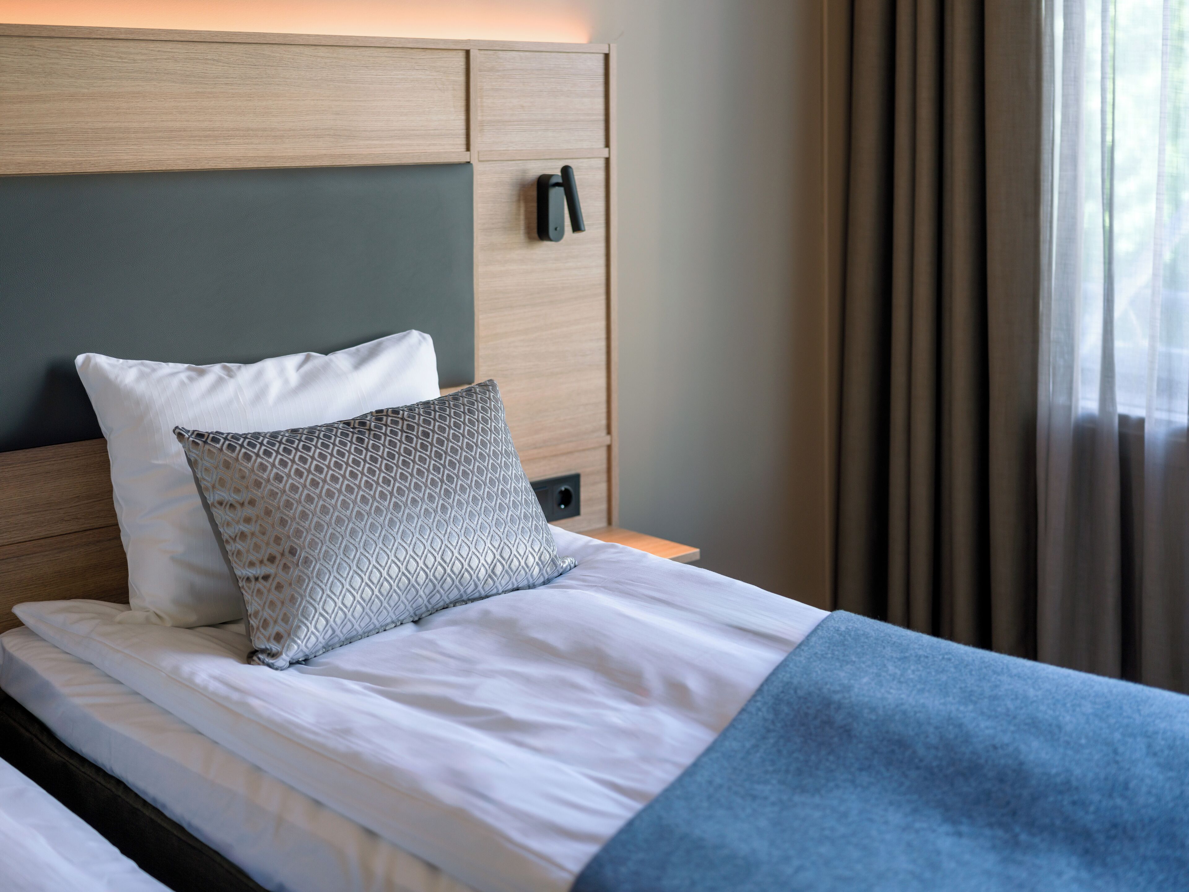 A neatly made bed with white linens and a blue blanket, a patterned pillow, and a wooden headboard with a built-in phone.